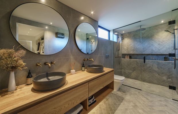Contemporary bathroom with a double floating timber vanity, two round vessel basins, wall-mounted bronze-toned tapware, large circular mirrors, and a spacious frameless glass shower enclosure. The room features dark grey textured wall tiles, coordinated floor tiles, warm timber joinery, recessed lighting, and a high horizontal window.
