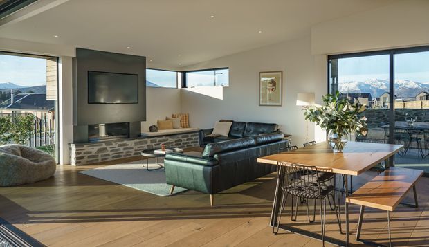Bright open-plan living and dining area with expansive glazing and mountain views. The room features wide-plank timber flooring, a stone-faced fireplace wall with built-in television, a dark leather sectional sofa, and a large timber dining table with wire-frame chairs and bench seating. Contemporary furnishings and abundant natural light create a warm, airy atmosphere.