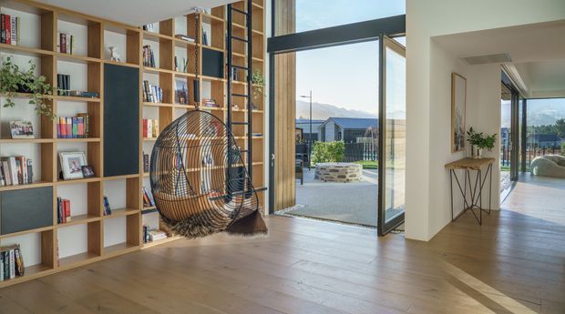 A bright, contemporary open-plan living area with extensive built-in timber shelving, integrated dark display panels, a hanging rattan egg chair, and wide timber flooring. Full-height glazing and an open glass door connect the room to an outdoor terrace, with mountain scenery visible beyond. A hallway and console table lead to another glazed living space.