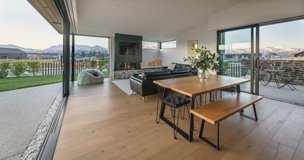 Open-plan contemporary living and dining area with full-height sliding glass doors opening to a terrace and lawn. The room features light timber flooring, a black-framed fireplace or media wall with stacked-stone detailing, a dark leather sectional sofa, and a timber dining table with bench seating. Mountain and suburban views are visible beyond the glazing.