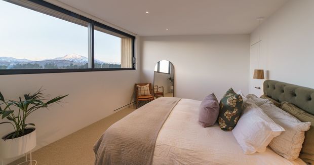 A bright, contemporary bedroom with a large corner window overlooking a mountainous landscape. The room features a low upholstered bed with layered neutral bedding and patterned cushions, a tufted green headboard, warm beige carpet, a potted plant, a timber lounge chair, and a freestanding arched mirror. Minimal recessed lighting and white walls create an airy, calm atmosphere.