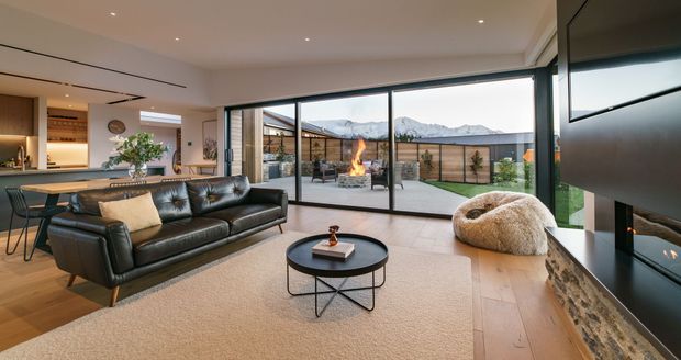 Contemporary open-plan living room with a black leather sofa, round coffee table, textured lounge chair, timber flooring and a large built-in fireplace feature wall. Floor-to-ceiling sliding glass doors open to a paved courtyard with an outdoor fire, with mountain scenery visible beyond.
