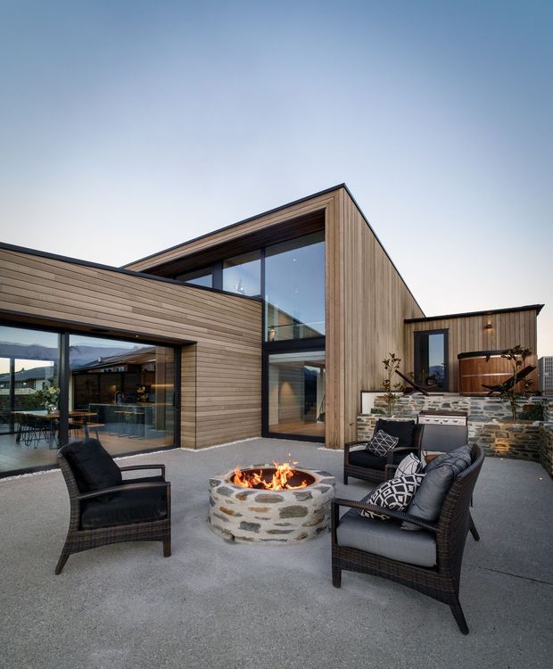 Contemporary multi-level home with extensive floor-to-ceiling glazing and warm horizontal timber cladding, arranged around a spacious paved outdoor terrace. The terrace features dark woven lounge chairs, a circular stone fire pit with an active fire, built-in stonework, and a raised outdoor spa or plunge pool area. Interior kitchen and dining spaces are visible through the glazing, with mountainous surroundings in the distance.