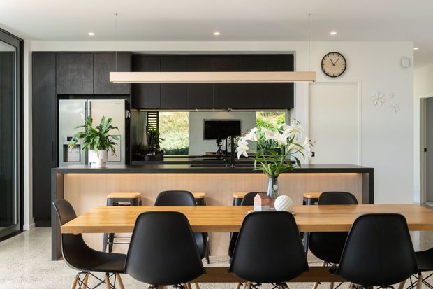 Contemporary open-plan kitchen and dining area with a large timber dining table and black dining chairs in the foreground. The kitchen features dark timber cabinetry, a light timber island with a dark benchtop, integrated appliances, a tiled or stone splashback, and a window overlooking greenery. White walls, recessed ceiling lights, and black-framed glazing create a clean, modern interior.