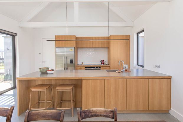 Bright, open-plan contemporary kitchen centred on a large timber island with integrated cabinetry, grey benchtops, bar seating, stainless-steel refrigerator, and a compact rear cooking zone. White walls and a pitched ceiling with exposed triangular structural framing create an airy, simple interior. Large windows and glazed doors bring in abundant natural light.