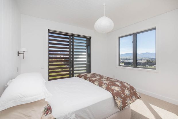 Bright, compact bedroom with a white upholstered bed, white bedding, and a brown floral-patterned throw. The room has white walls and ceiling, pale carpet, dark-framed windows, and exterior horizontal louvred screening. A white spherical pendant light hangs centrally, while mountain scenery is visible through the windows.