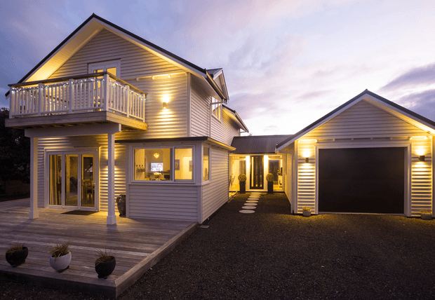 Exterior dusk view of a substantial two-storey coastal-style home with light horizontal weatherboard cladding, a pitched gabled roof, covered entry, upper-level balcony, timber deck, and an attached single-storey garage. Warm architectural lighting illuminates the facade, deck, and central passage leading to the entrance.