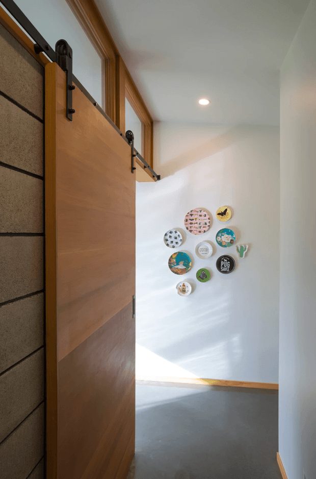 A narrow, contemporary interior passage viewed through a partially open sliding timber door. The door uses exposed dark metal barn-door hardware and sits beside a textured dark wall. White walls, grey flooring, timber trim, recessed ceiling lighting, and a small arrangement of colourful circular artworks create a bright, playful hallway or landing.