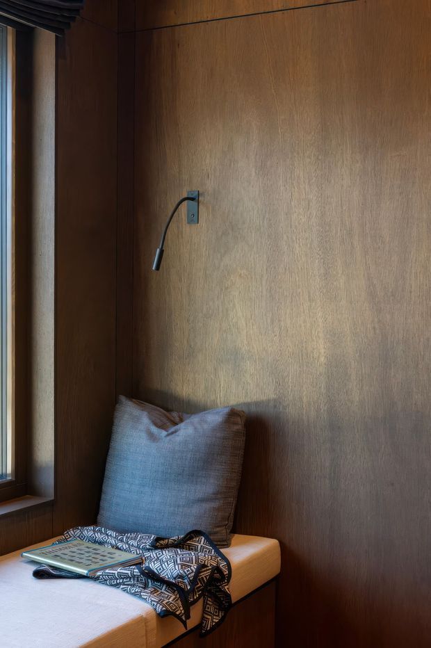 A compact built-in window seat or reading nook, clad in warm dark timber panelling. The seat has a tan upholstered cushion, a large muted blue-grey pillow, a patterned throw, and a small wall-mounted reading light. A narrow window with a dark timber frame is visible at left. The image has a warm, intimate, refined interior character with strong timber detailing.