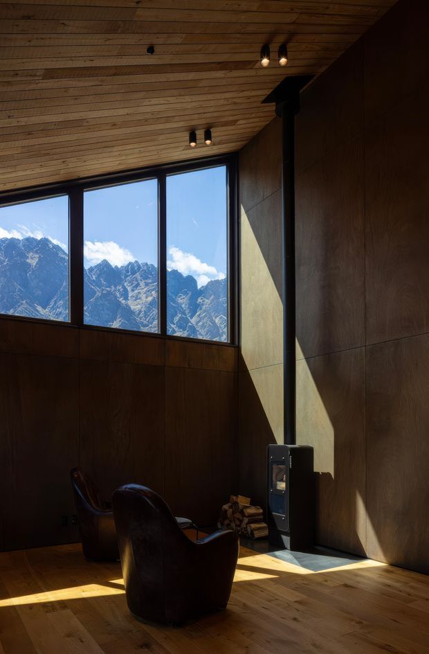 A dramatic, double-height mountain retreat interior with dark timber wall panelling, a warm timber-lined sloping ceiling, and expansive black-framed windows overlooking rugged alpine peaks. Two dark leather lounge chairs sit beside a compact black wood-burning stove and stacked firewood. Strong sunlight creates angular shadows across the walls and timber floor, giving the room a quiet, moody cabin atmosphere.
