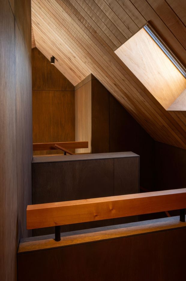 Interior architectural view of a warm, compact stairway or landing with extensive dark timber wall panelling and cabinetry, a timber balustrade, and a sloped timber-lined ceiling. A linear skylight introduces natural light from above, highlighting the angular roof geometry and rich wood grain.