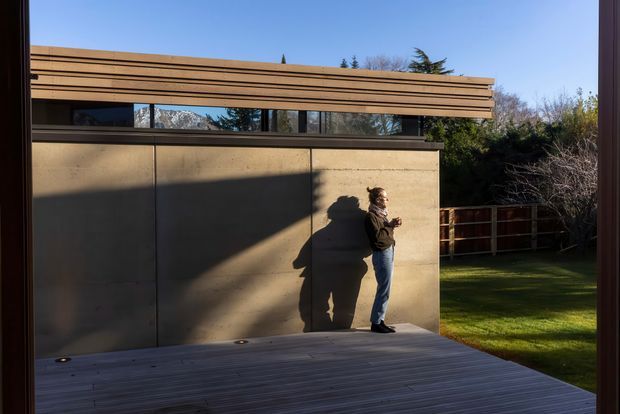 Exterior architectural view of a compact modern structure with a long horizontal timber-clad roof or screen, narrow clerestory glazing, and a smooth warm-toned concrete wall. A timber deck occupies the foreground, with a fenced grassy garden beyond. Strong late-afternoon sunlight creates dramatic shadows across the wall.