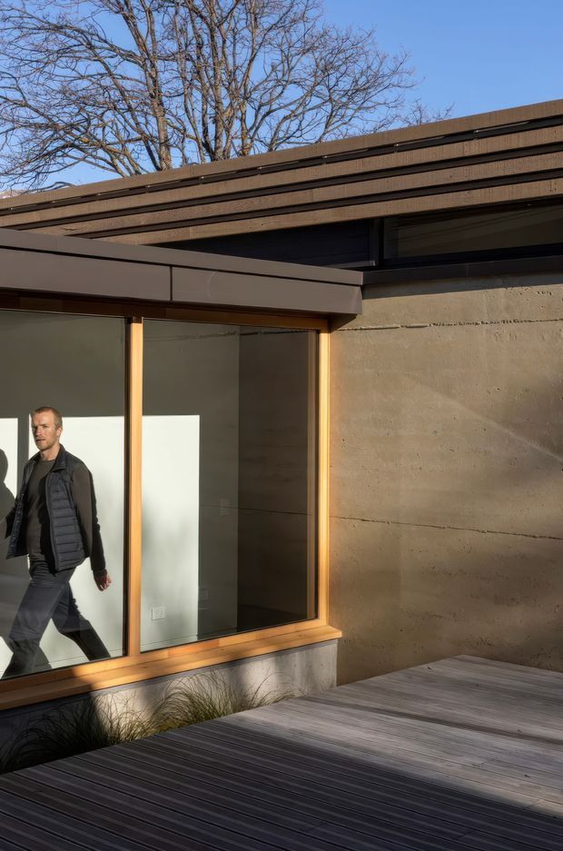 Exterior architectural photograph of a modern low-profile building opening onto a timber deck. The facade combines rough exposed concrete walls, warm timber-framed glazing, dark metal-framed clerestory windows, and layered horizontal timber roof cladding. A leafless tree and a person are visible, but the focus is the building envelope and outdoor deck.