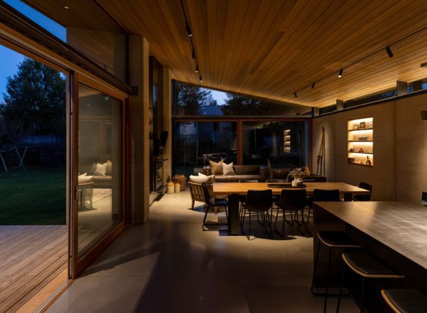 A warm, open-plan residential interior combining a dining area, living room, and kitchen. Full-height timber-framed glazing opens onto a timber deck and garden, while a timber-lined ceiling extends across the space. The room features dark built-in cabinetry, an illuminated display niche, a dining table with black chairs, a lounge seating area, and a kitchen island with stools. Track lighting and concealed warm lighting create an intimate evening atmosphere.
