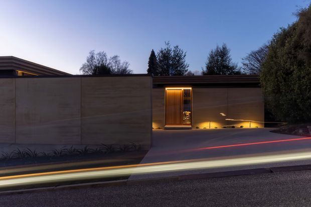 A dusk exterior view of a low, minimalist residential property behind a tall smooth concrete boundary wall. A recessed timber entry door is warmly illuminated, with linear wall-washing lights along the driveway and subtle horizontal roof detailing. Long-exposure light trails from passing traffic appear in the foreground, while mature vegetation frames the right side.
