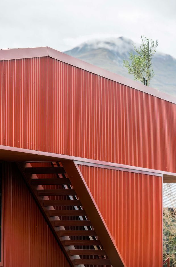 Close-up exterior architectural photograph of a contemporary building clad in vertically ribbed red corrugated metal. An exposed external metal stair climbs along the facade beneath a projecting upper volume. A sloping roofline, sparse tree, and snow-dusted mountain are visible in the background, suggesting a mountainous setting.