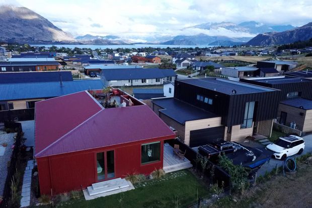 Aerial view of a contemporary residential neighbourhood beside a broad lake, surrounded by steep mountainous terrain. The foreground shows modern homes with dark vertical cladding, timber accents, and vivid red and blue metal pitched roofs, along with driveways and small landscaped yards.