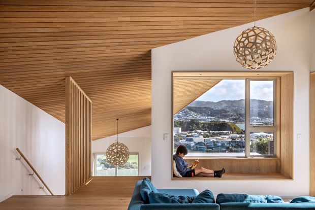 A warm contemporary interior with a slatted timber ceiling, white walls, and a large timber-framed window seat overlooking a mountainous urban landscape. A blue sectional sofa occupies the foreground, while spherical woven pendant lights and a vertical timber privacy screen add architectural detail. A person is seated in the window nook.