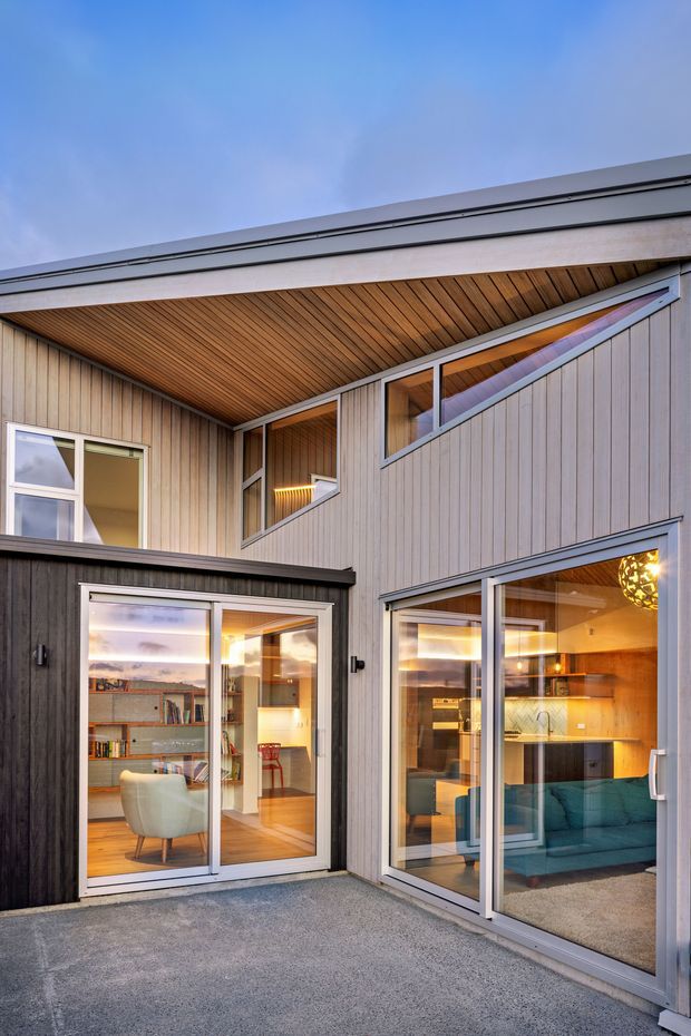 Exterior view into a compact contemporary courtyard, showing a two-storey residential facade clad in vertically boarded timber. The recessed courtyard has a warm timber-lined soffit, broad glazed sliding doors and windows, and visible interiors including a study or living space on the left and a kitchen/living area on the right. Grey concrete paving and dark grey window and door frames create a restrained modern palette, with warm interior lighting visible through the glazing.