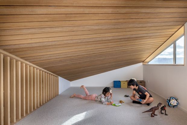 A carpeted attic-style playroom beneath a dramatically sloping timber-lined ceiling. A low vertical timber balustrade divides the space, while a triangular window brings in daylight and views outside. Children are playing on the floor among toys and storage boxes.