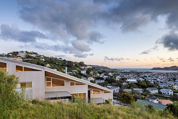 Exterior view of a modern, low-profile house set on a steep grassy hillside, with pale vertical cladding, broad angular glazing, and a sloping roof. A dense coastal suburban settlement and open water are visible beyond, under a dramatic cloudy sky.