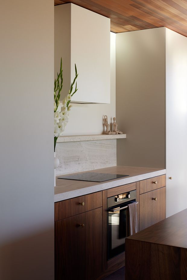 A compact contemporary kitchen with dark timber cabinetry, integrated oven, induction cooktop, pale stone benchtops and splashback, and warm timber lining on the ceiling. Sculptural decor and flowers add understated character, while a partial island or dining surface appears in the foreground.