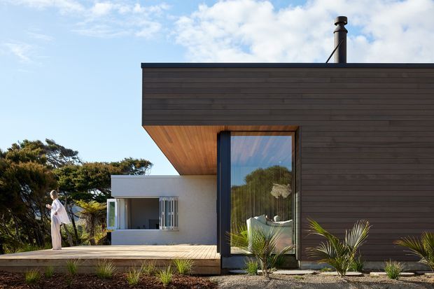 Exterior view of a contemporary single-storey home with a dark horizontal timber-clad box-like volume, broad cantilevered roof/eave, large glazed opening, and a lighter rendered pavilion behind. A paved terrace and sparse coastal-style planting sit in the foreground, with mature trees in the background.