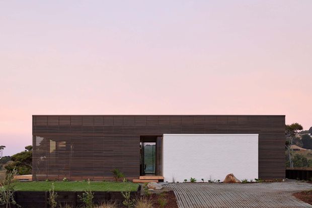 Single-storey contemporary residence viewed from the front, with a long rectangular form, dark horizontal timber-like screening across the facade, a contrasting white rendered wall, recessed glazed entry, and a broad gravel driveway. Low landscaping and a soft pink sky surround the minimalist building.