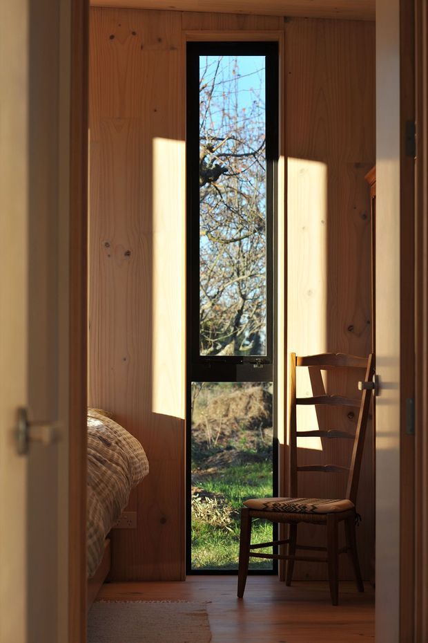 A warmly lit, narrow bedroom or sleeping nook lined with vertical natural timber panelling. A tall black-framed glazed door or window provides a view to trees and garden beyond, while a wooden ladder-back chair sits in the sunlight beside it. Part of a bed with woven or patterned bedding is visible at left, and timber flooring continues through the space.