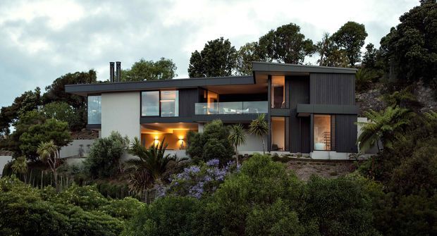 Exterior view of a contemporary multi-level residence set within dense landscaped greenery. The house has a strong horizontal composition, dark flat roof planes, dark vertical cladding, pale rendered wall sections, extensive glazing, recessed warm lighting, and a glass-balustraded upper terrace.