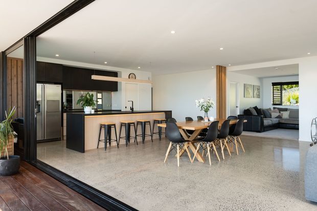 Open-plan contemporary kitchen, dining, and living area viewed from a timber deck through large black-framed sliding glazing. The space features a dark kitchen with a light island and breakfast bar, terrazzo-style flooring, a timber dining table with black chairs, and a partially visible lounge area beyond.