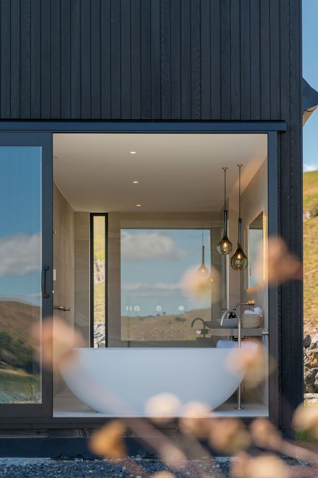 A modern bathroom viewed through a large glazed opening, featuring a freestanding white bathtub, wall-mounted basin with curved tapware, pale stone or tile walls, dark timber cladding and framing, pendant lights, and expansive glazing reflecting a rugged coastal or hillside landscape. The foreground contains softly blurred vegetation.