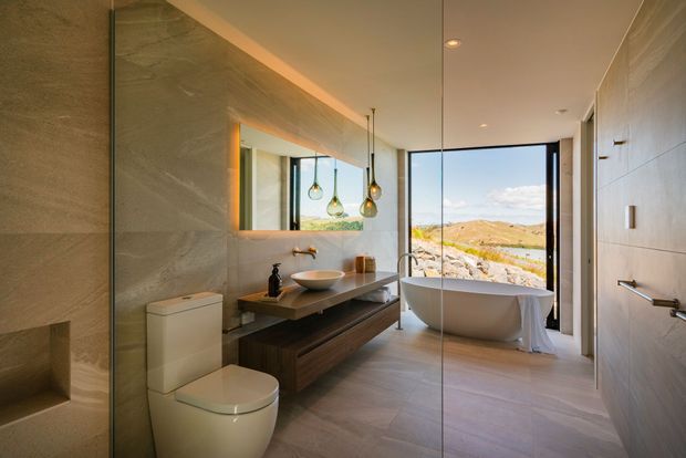 Contemporary bathroom with a freestanding oval bathtub positioned beside full-height glazing, a wall-mounted timber vanity with vessel basin, toilet, large mirror, recessed shower niche, and warm pendant lighting. Large-format stone-look wall and floor finishes create a refined, natural-toned interior, with a rocky sunlit landscape visible beyond the window.