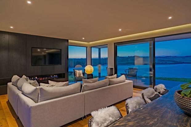 A refined contemporary living room with large corner glazing overlooking a blue waterfront landscape. The room features a substantial pale sectional sofa, dark timber wall panelling with a wall-mounted television and linear fireplace, recessed ceiling lights, warm concealed lighting, timber flooring, and a dark dining table with fur-covered chairs in the foreground.
