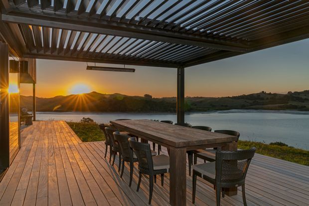A covered outdoor dining terrace overlooking a calm body of water at sunset. The space features a large timber dining table with woven chairs, timber decking, dark structural posts, and a slatted pergola roof with integrated lighting. Rolling hills and the setting sun form the background.