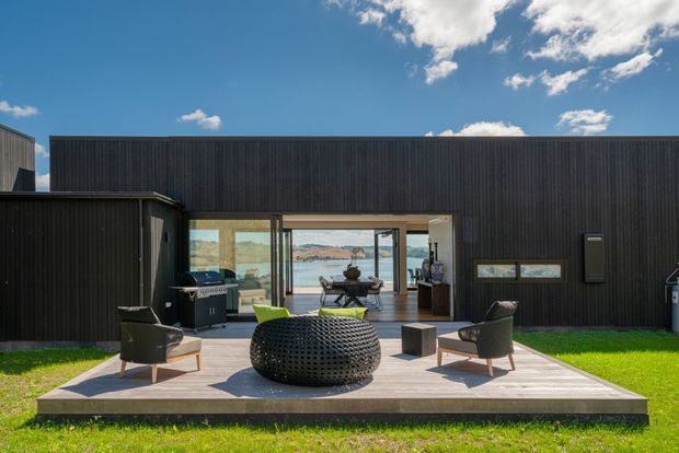Contemporary dark-clad single-storey residence with a broad glazed opening connecting an outdoor timber deck to a bright interior and a visible waterfront landscape beyond. The deck includes an outdoor seating arrangement and barbecue, while the building has a simple low-profile rectangular form with vertical ribbed cladding.
