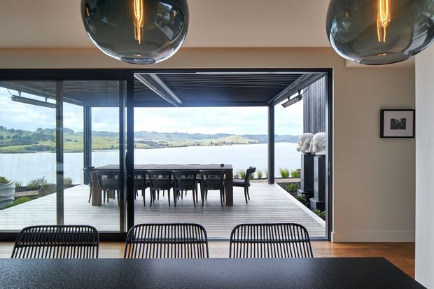 A contemporary dining area opens through large glazed sliding doors onto a covered timber deck overlooking a broad lake and rolling green hills. A long dark dining table and woven black dining chairs occupy the foreground, with spherical pendant lights overhead. The space features expansive glazing, a slatted ceiling and screened outdoor dining terrace.