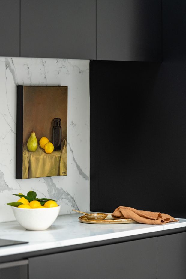 Close-up view of a sophisticated modern kitchen featuring dark matte cabinetry, a dramatic black wall panel, and a white marble-look splashback and benchtop. A bowl of lemons, plate, napkin, and framed still-life artwork add warmth and styling to the otherwise monochromatic space.