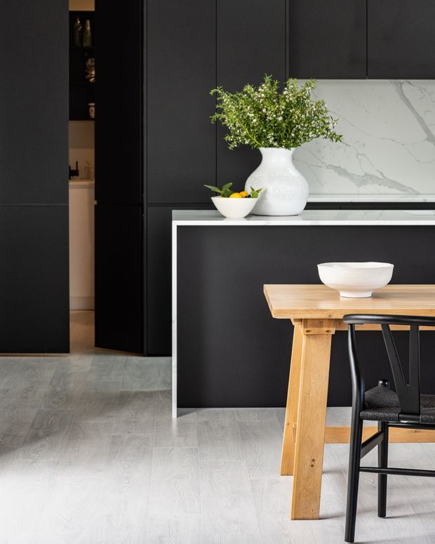 A refined contemporary kitchen and dining area featuring matte black full-height cabinetry, a black kitchen island with a slim white edge, and a white marble-look splashback. A light timber dining table with black chairs occupies the foreground, while a white ceramic vase and bowl provide understated styling. Pale timber-look flooring contrasts with the dark cabinetry.