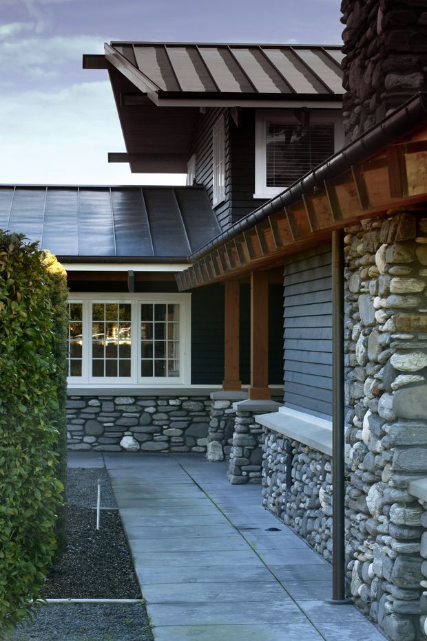 Exterior architectural view of a compact, multi-level home surrounding a paved entry courtyard. The building combines dark horizontal weatherboard siding, extensive rounded natural-stone masonry, white-trimmed multi-pane windows and doors, and standing-seam metal roofs with pronounced eaves. A covered walkway and timber-supported overhang frame the entry, creating a sheltered, textural approach.