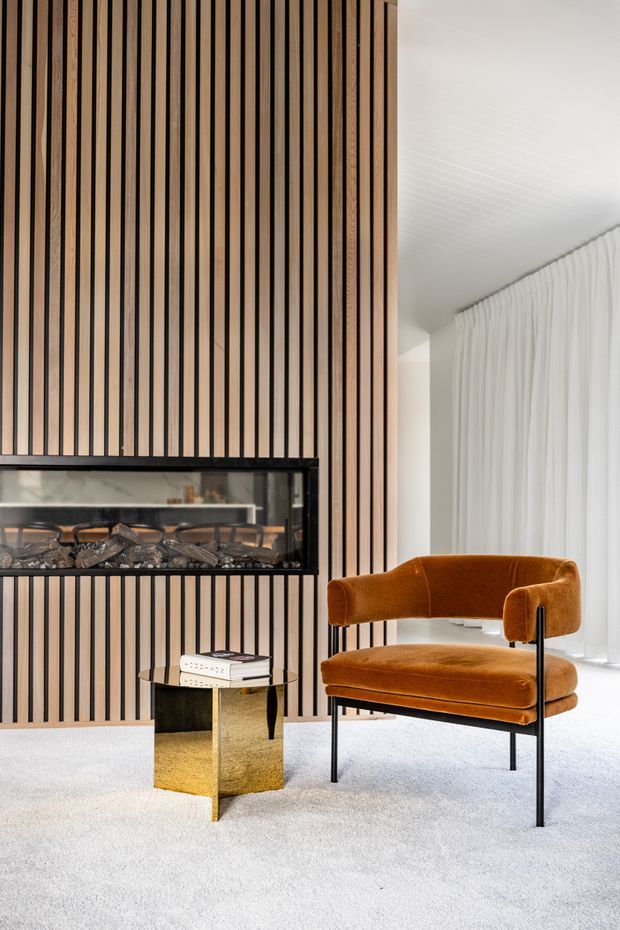 A refined living room featuring a full-height vertical timber-slat feature wall with an inset linear fireplace, a burnt-orange velvet lounge chair, a small metallic side table, white sheer curtains, and pale carpet. The space combines warm natural wood, soft neutral finishes, and sculptural contemporary furniture.