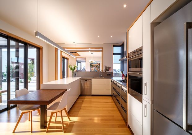 Bright, open-plan contemporary kitchen with extensive white built-in cabinetry, integrated stainless-steel appliances, a long central island, and warm timber flooring. A compact timber dining table with white chairs sits beside full-height glazed doors, while timber-framed openings, exposed timber ceiling detailing, and generous daylight create a warm, refined interior.