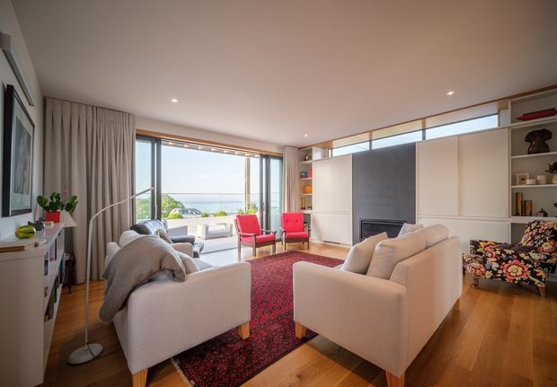 Bright, warmly furnished living room with polished timber flooring, two cream armchairs and a cream sofa arranged around a red patterned rug and a built-in fireplace. Full-height glazing and sliding doors open toward a balcony or terrace with a distant coastal outlook. Built-in shelving and cabinetry line the fireplace wall, while curtains, artwork, plants, and colourful occasional chairs add character.