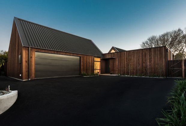 A contemporary residential exterior featuring a detached-looking garage or outbuilding with a steep gabled roof, dark standing-seam metal roofing, vertical timber cladding, and a large concealed-panel garage door. A matching timber-clad boundary wall and gated entry enclose a broad dark paved driveway, with subtle planting along the edges.