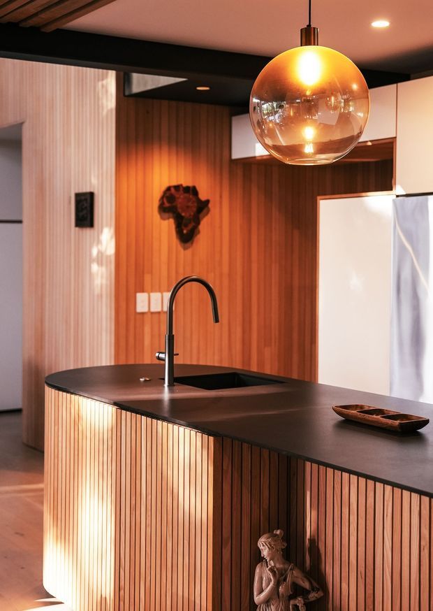 A warm, moody kitchen interior centred on a large curved island with a dark smooth benchtop, integrated sink and matte black gooseneck tap. The island and rear wall feature vertical timber slat panelling, complemented by white upper cabinetry and a large white refrigerator. A glowing amber glass pendant hangs above the island, while black ceiling beams and timber ceiling boards add strong architectural contrast. The space includes eclectic decorative elements, including a small wall-mounted sculpture and a figurine at the island base.
