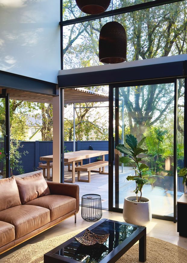 A bright, contemporary indoor-outdoor living area with floor-to-ceiling black-framed glazing opening onto a covered timber dining deck. The interior features a brown leather sofa, black glass coffee table, woven rug, potted plant, and sculptural pendant lights. Outside, a timber pergola, blue privacy fencing, and a substantial leafy tree are visible.