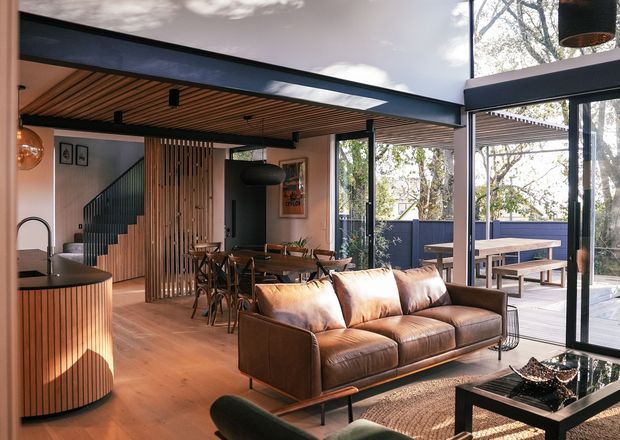 Open-plan contemporary living and dining area with a compact kitchen, warm timber ceiling and flooring, vertical timber screening, black structural framing, and expansive glazed doors opening to an outdoor terrace. A brown leather sofa faces a low coffee table, while a dining table sits behind it near the stairway. The space has abundant natural light and a strong indoor-outdoor connection.