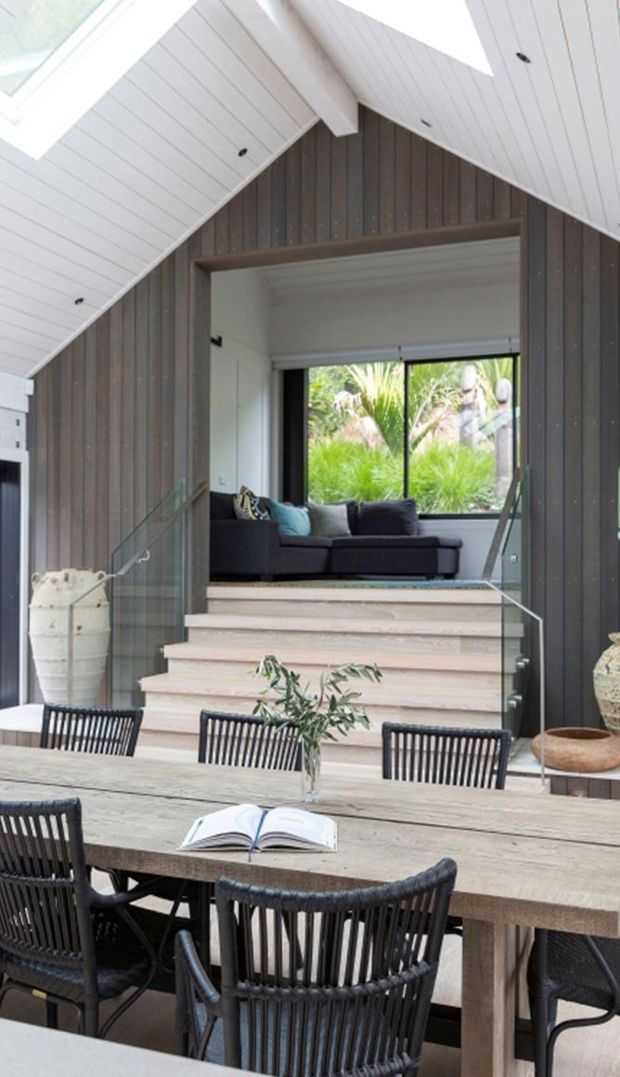 Bright contemporary dining area with a large timber dining table and black woven dining chairs, opening toward a raised lounge nook. The space features dark vertical timber wall cladding, pale timber stairs, extensive glazing, and a dramatic white pitched ceiling with exposed beam detailing. Lush planting is visible beyond the window.