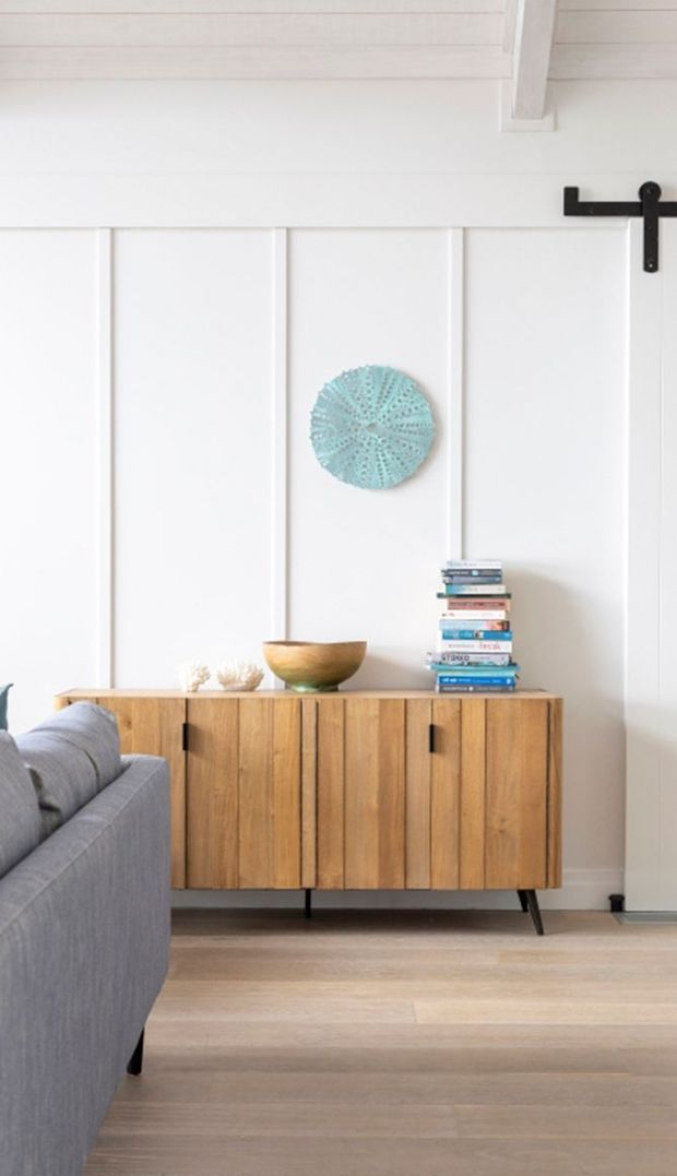 A bright, neatly styled living room corner featuring white vertical wall panelling, exposed white ceiling boards and a black sliding barn-door track. A warm timber sideboard with vertical cabinet fronts sits against the wall, topped with books and a wooden bowl. A grey upholstered sofa occupies the foreground, with pale timber flooring and a turquoise circular wall accent adding colour.