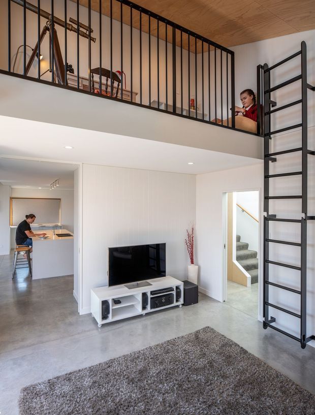 Bright contemporary double-height living room with polished concrete flooring, white walls and built-in cabinetry, a black metal mezzanine balustrade and ladder, and a timber-lined ceiling. A compact kitchen is visible to the left, with a stairway leading upward at the rear.